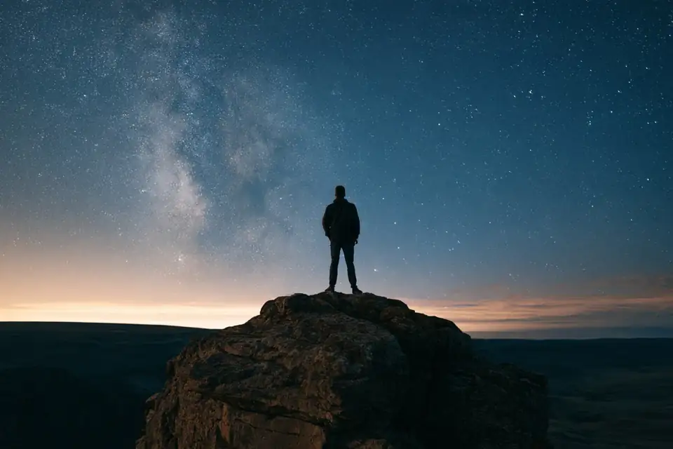 un homme au bord d’une falaise, face à un ciel étoilé immense.