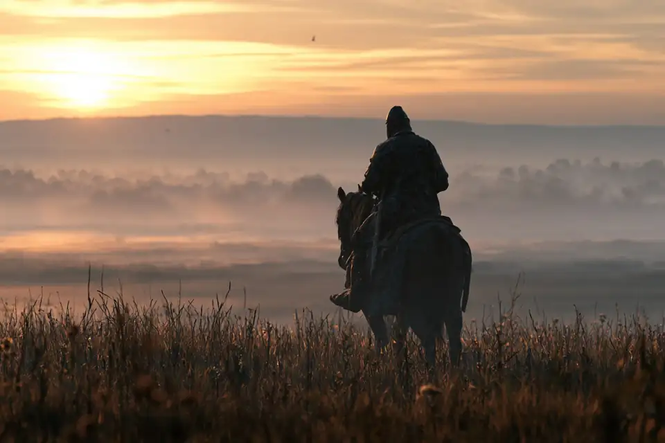 un cavalier en armure sur un cheval nerveux, face à une plaine couverte de brume à l’aube.