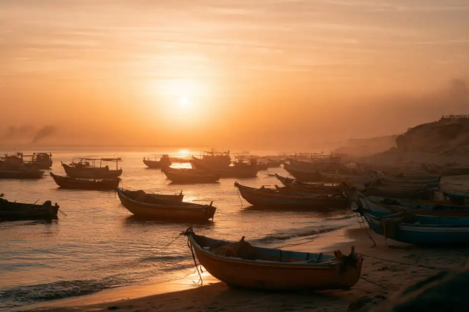 une plage à l’aube couverte de petits bateaux, fumée à l’horizon.